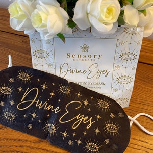 May include: A black eye mask with gold eye and star motifs and the text Divine Eyes, positioned in front of a white product packet labelled Sensory Retreats Divine Eyes Heating Eye Mask with lavender scent, featuring a bouquet of white roses.