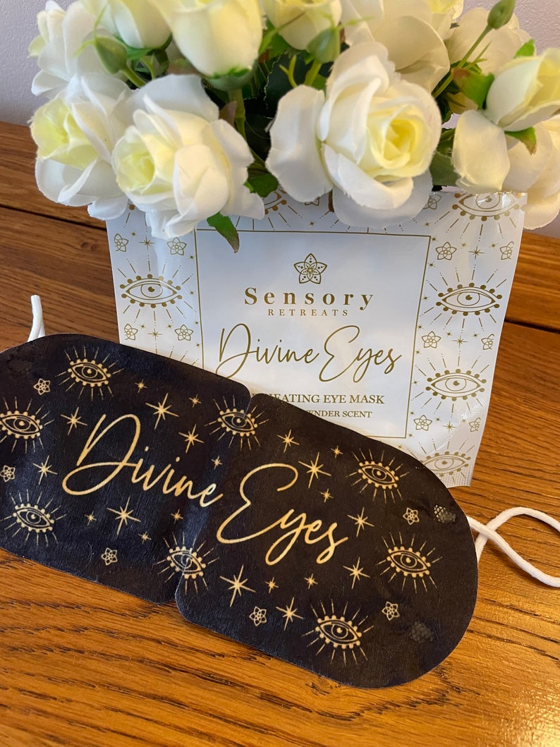 May include: A black eye mask with gold eye and star motifs and the text Divine Eyes, positioned in front of a white product packet labelled Sensory Retreats Divine Eyes Heating Eye Mask with lavender scent, featuring a bouquet of white roses.