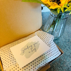 May include: An open cardboard shipping box containing white tissue paper with a black polka dot pattern. A rectangular white card rests on top of the tissue paper, featuring the text "Sending you a hug" in black script over a sage green brushstroke.