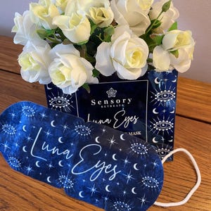 May include: A blue sleep mask with the words "Luna Eyes" in white script, adorned with crescent moons, stars, and eye symbols. The mask is placed in front of a box with the words "Sensory Retreats" and a bouquet of white roses.