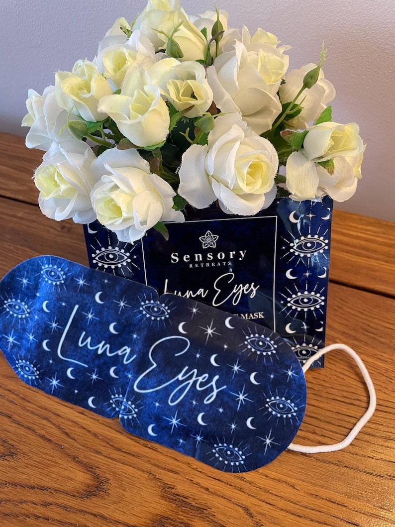 May include: A blue sleep mask with the words "Luna Eyes" in white script, adorned with crescent moons, stars, and eye symbols. The mask is placed in front of a box with the words "Sensory Retreats" and a bouquet of white roses.