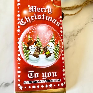 May include: A rectangular milk chocolate bar in a red wrapper featuring a snow globe graphic with two snowmen holding chocolate bars. The text reads Merry Christmas To you SOLID MILK CHOCOLATE BAR and Hames. The wrapper has white star accents.