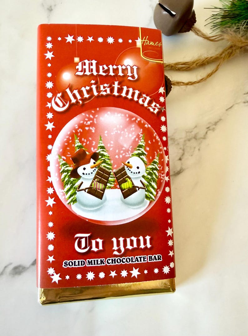 May include: A rectangular milk chocolate bar in a red wrapper featuring a snow globe graphic with two snowmen holding chocolate bars. The text reads Merry Christmas To you SOLID MILK CHOCOLATE BAR and Hames. The wrapper has white star accents.