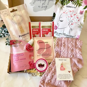 May include: A gift box containing a card reading Sending you a hug, Ask Mummy & Daddy Prosecco gummies, two Twinings Glow tea bags, a DermaV10 hair mask, Face Facts rose jelly mask, Grace & Stella eye masks, a rose shower steamer, and pink socks with a heart-shaped pocket hug charm.