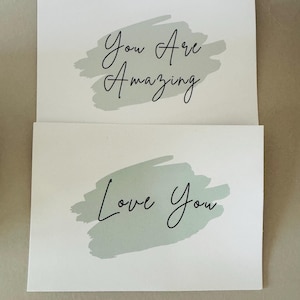 May include: Two rectangular white cards featuring sage green brushstroke accents. The top card displays the black text "You Are Amazing" in a cursive font, while the bottom card features the black text "Love You" in the same cursive style.