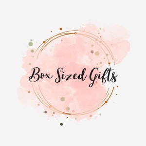 May include: A logo featuring the text "Box Sized Gifts" in elegant script. The text is set against a soft pink watercolor background, encircled by a double gold ring. Small gold and green speckles add detail.