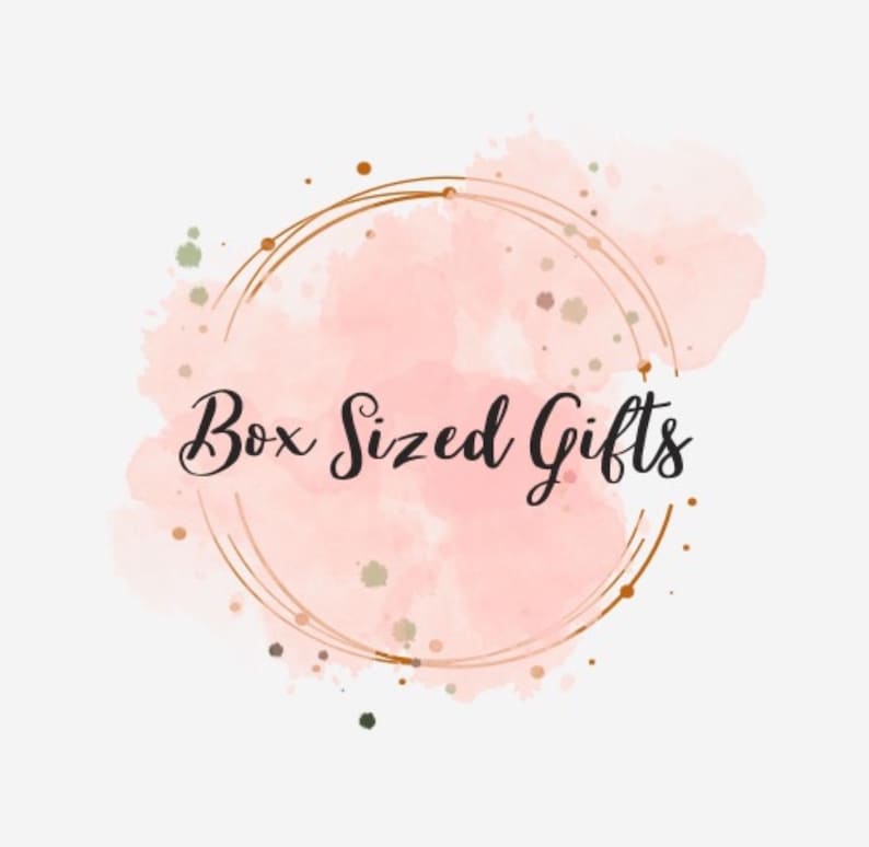 May include: A logo featuring the text "Box Sized Gifts" in elegant script. The text is set against a soft pink watercolor background, encircled by a double gold ring. Small gold and green speckles add detail.