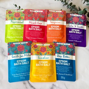 May include: Seven colourful 35g pouches of Epsom bath salt arranged in a group. Each pouch features a floral pattern at the top and text including Peppermint, Muscle Ease, Tangerine, Calm & Soothe, Smile for Miles, Sunshine, and Sleep Better.
