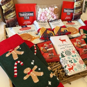 May include: A gift box containing Galaxy hot chocolate packets, Yorkshire Tea, marshmallows, Walkers shortbread, Daelmans stroopwafels, a chocolate bar with a reindeer design, and green socks featuring gingerbread men and candy canes.