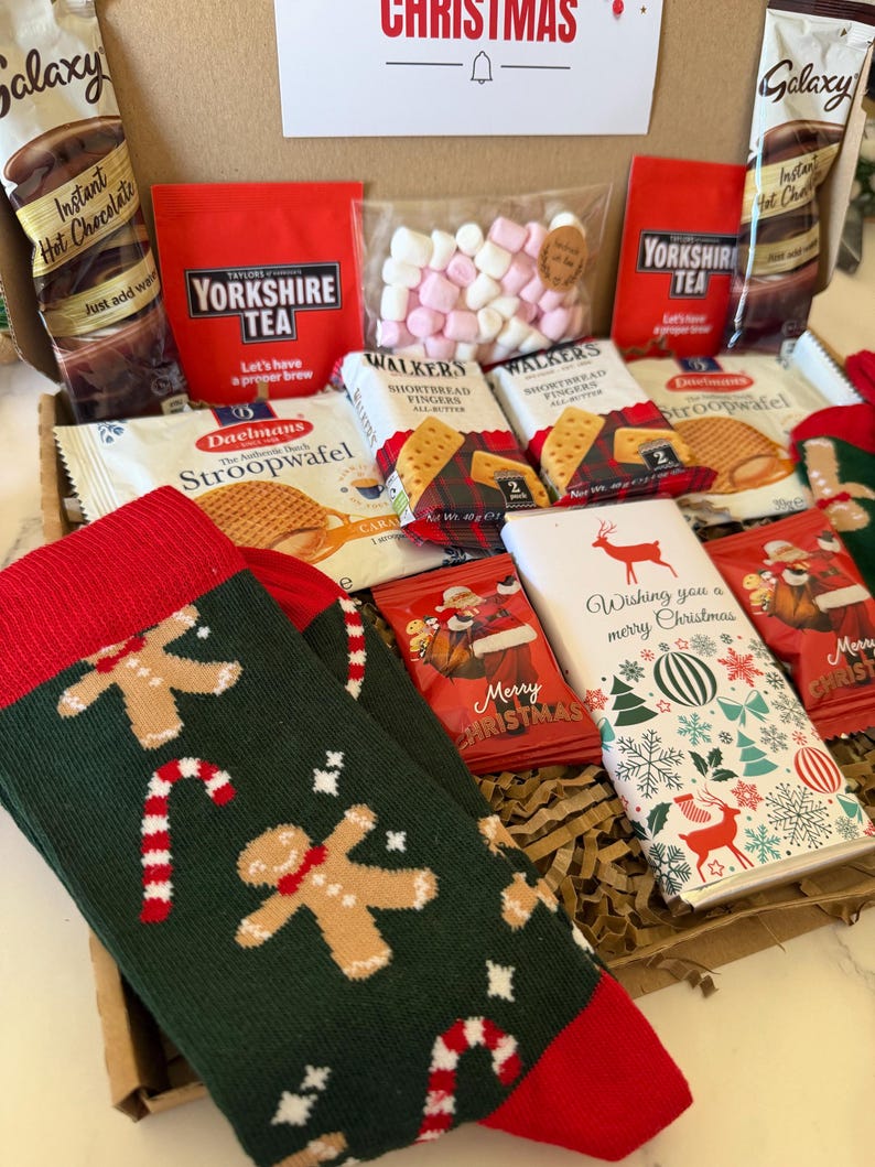 May include: A gift box containing Galaxy hot chocolate packets, Yorkshire Tea, marshmallows, Walkers shortbread, Daelmans stroopwafels, a chocolate bar with a reindeer design, and green socks featuring gingerbread men and candy canes.