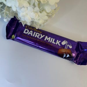 May include: A purple Cadbury Dairy Milk chocolate bar with white text and a gold logo. The wrapper has a shiny, metallic finish. The bar is positioned near a bouquet of white flowers.