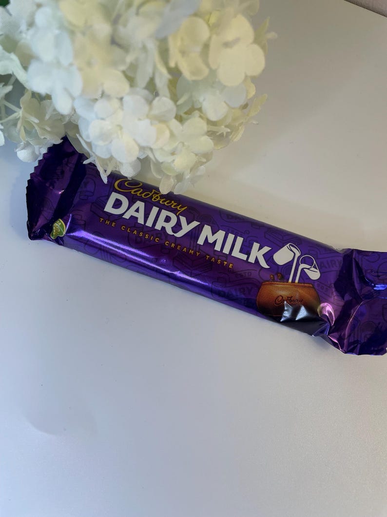 May include: A purple Cadbury Dairy Milk chocolate bar with white text and a gold logo. The wrapper has a shiny, metallic finish. The bar is positioned near a bouquet of white flowers.