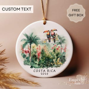 May include: A round ceramic ornament with a watercolor design featuring two toucans perched on a branch amidst tropical foliage. The ornament has the text "COSTA RICA 2025" and "CUSTOM TEXT" at the top. A beige string is attached for hanging.