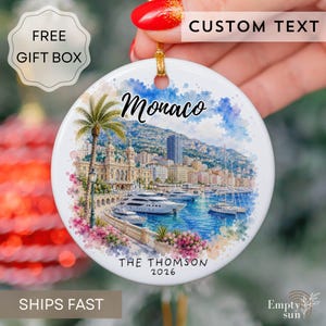 May include: A round white ceramic ornament featuring a watercolor illustration of the Monaco harbor with yachts, buildings, and palm trees. The text "Monaco" appears at the top, and "THE THOMSON 2026" is printed at the bottom in black lettering.
