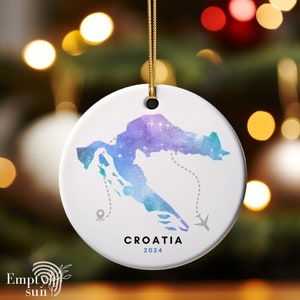 May include: A white ceramic ornament with a watercolor map of Croatia in shades of blue, purple, and green. The ornament features a plane and a pin, and the text "CROATIA 2024".