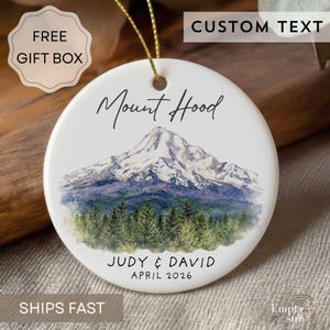 May include: A round white ceramic ornament featuring a watercolor illustration of Mount Hood with a forest at the base. The text reads Mount Hood, Judy & David, and April 2026. A gold string is attached to the top for hanging the ornament.