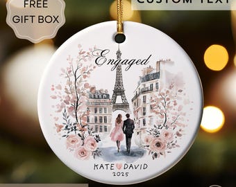 May include: A white ceramic ornament with a gold string, featuring a watercolor illustration of a couple walking in Paris, with the Eiffel Tower in the background. The ornament says "Engaged" and includes the names "Kate & David" and the year "2025".