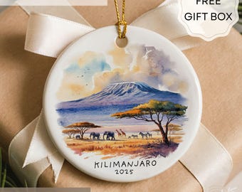 May include: A round ceramic ornament featuring a watercolor painting of Mount Kilimanjaro and African wildlife. The ornament has a gold string for hanging and the text "KILIMANJARO 2025". The background is a soft, neutral color.