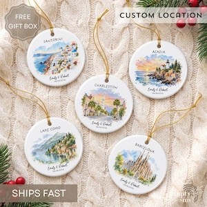 May include: Five round white ceramic ornaments with gold hanging strings. Each features a unique watercolor illustration of a travel destination, including Santorini, Acadia, Charleston, Lake Como, and Barcelona, with custom names and dates printed below.