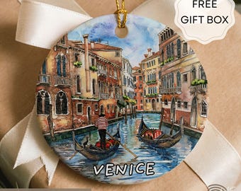 May include: Round ceramic ornament featuring a watercolor painting of Venice, Italy. The artwork depicts gondolas on a canal with buildings on either side. The word "VENICE" is written across the bottom. A gold string is attached for hanging.