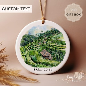 May include: A round white ceramic ornament with a watercolor painting of a Bali landscape. The image features green rice terraces, palm trees, and small buildings. The text "BALI, 2025" is printed at the bottom. A beige string is attached for hanging.