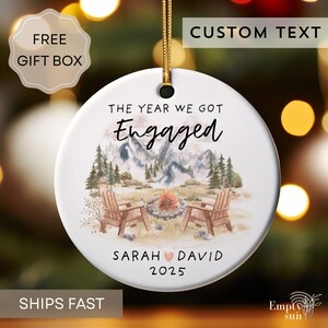 May include: White ceramic ornament with a gold string, featuring a watercolor illustration of a mountain scene with two chairs and a campfire. Text reads "THE YEAR WE GOT Engaged" and names "SARAH DAVID" with the year "2025".