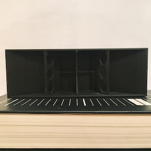 Könnte beinhalten: Ein schwarzer Lautsprechergehäuse mit mehreren Anschlüssen an der Vorderseite. Der Lautsprecher steht auf einem schwarzen Buch mit einem weißen Rücken.