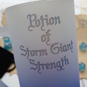 Może przedstawiać: Kubek z matowego szkła z niebieskim efektem ombre. Kubek ma uchwyt i napis "Potion of Storm Giant Strength" w ozdobnym kroju pisma.