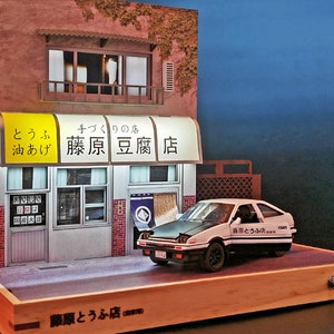 May include: A miniature model of a Japanese tofu shop with a white Toyota AE86 car parked in front. The shop has a yellow sign that reads "とうふ 油あげ 藤原豆腐店" which translates to "Tofu, Oil-fried Tofu, Fujiwara Tofu Shop". The car has a black and white sticker that reads "藤原とうふ店" which translates to "Fujiwara Tofu Shop".