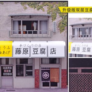 May include: A model of a Japanese tofu shop with a yellow awning that reads "とうふ 油あげ 藤原豆腐店" (tofu, oil-fried tofu, Fujiwara Tofu Shop). The shop has a red brick facade and a traditional Japanese door with a black and white design.