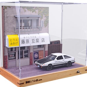 May include: A white Toyota AE86 with a black roof and black accents, including the words "藤原とうふ店" on the side, is parked in front of a small, white building with a yellow awning that reads "藤原豆腐店" and "とうふ 油あげ". The building is in a glass display case with a wooden base.