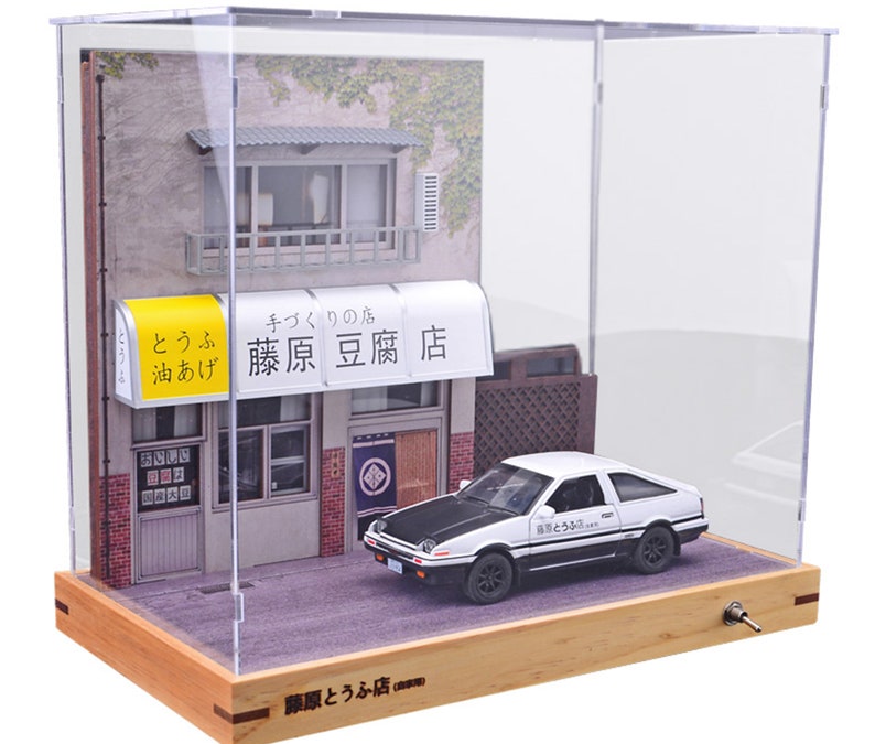 May include: A white Toyota AE86 with a black roof and black accents, including the words "藤原とうふ店" on the side, is parked in front of a small, white building with a yellow awning that reads "藤原豆腐店" and "とうふ 油あげ". The building is in a glass display case with a wooden base.