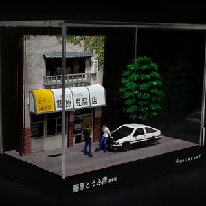 May include: A miniature diorama of a Japanese tofu shop with a white car parked in front. The shop has a yellow awning with the text "藤原豆腐店" in Japanese. The diorama is displayed in a clear acrylic case with a black base.