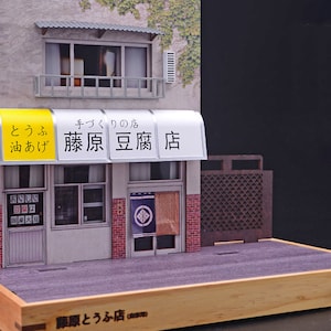 May include: A miniature model of a Japanese tofu shop with a yellow and white awning that reads "とうふ 油あげ 藤原豆腐店" (tofu, oil-fried tofu, Fujiwara Tofu Shop). The shop has a wooden door and two windows. The model is set on a wooden base with a switch.