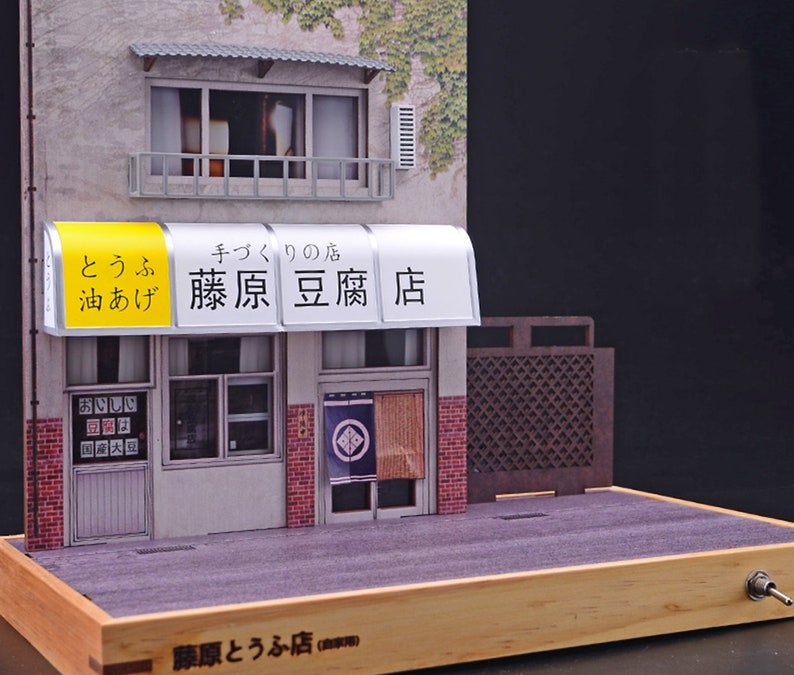 May include: A miniature model of a Japanese tofu shop with a yellow and white awning that reads "とうふ 油あげ 藤原豆腐店" (tofu, oil-fried tofu, Fujiwara Tofu Shop). The shop has a wooden door and two windows. The model is set on a wooden base with a switch.