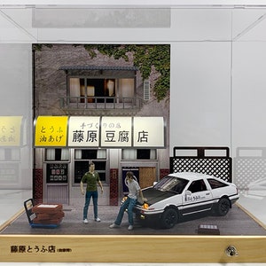 May include: A white diecast model car with black accents and the text "藤原とうふ店" on the side, sitting in front of a miniature storefront with a yellow awning that reads "藤原豆腐店". The storefront is made of wood and has a brick facade. Two miniature figures stand in front of the store.