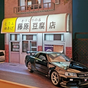 May include: A black Nissan Skyline GT-R R34 model car parked in front of a Japanese restaurant with a yellow awning that reads "と う ふ 油 あげ  藤原 豆腐 店" which translates to "Tofu Oil Fried  Fujiwara Tofu Shop".