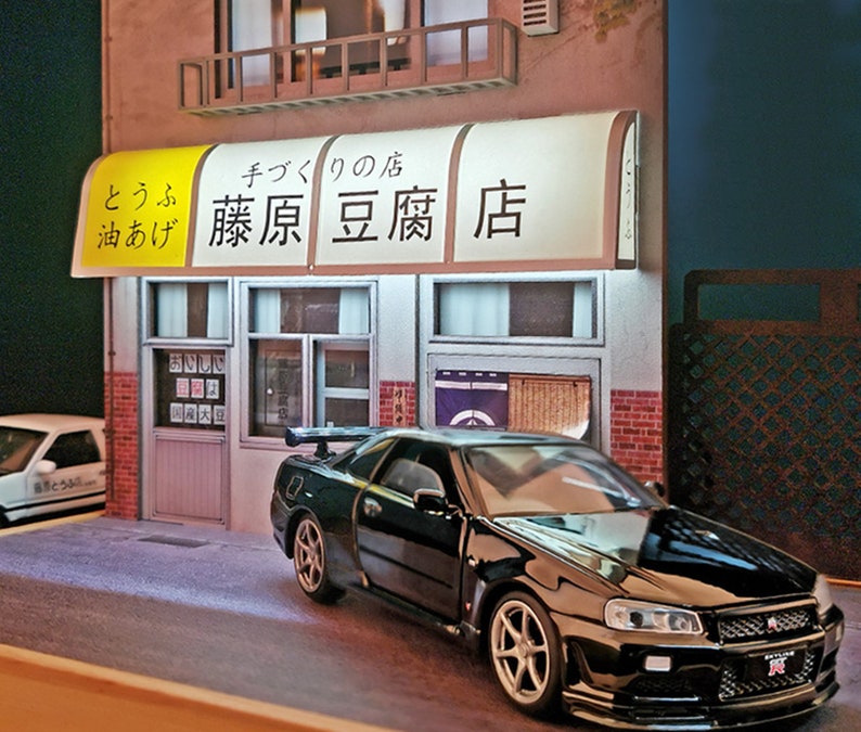 May include: A black Nissan Skyline GT-R R34 model car parked in front of a Japanese restaurant with a yellow awning that reads "と う ふ 油 あげ  藤原 豆腐 店" which translates to "Tofu Oil Fried  Fujiwara Tofu Shop".