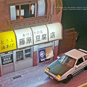 May include: A white toy car with a black hood and a Japanese logo on the side. The car is parked in front of a small, detailed model of a Japanese storefront with a yellow awning that reads "とうふ 油あげ 藤原豆腐店" in Japanese.