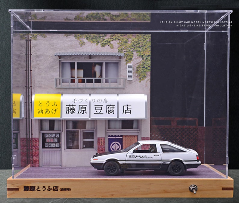 May include: A white diecast model car with the words "藤原とうふ店" on the side, sitting in a glass display case with a miniature storefront that says "藤原豆腐店" in Japanese. The storefront has a yellow sign that says "とうふ 油あげ" in Japanese.