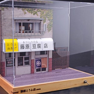 May include: A miniature model of a Japanese storefront with a sign that reads "キクノの店 藤原豆腐店" (Kikuno's Shop Fujiwara Tofu Store). The storefront is made of paper and wood and is displayed in a clear acrylic case with a wooden base.