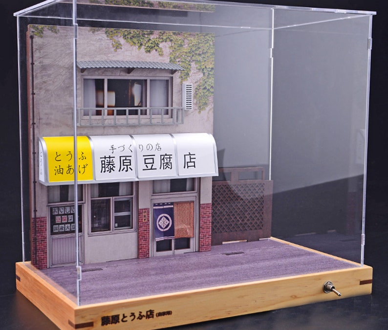 May include: A miniature model of a Japanese storefront with a sign that reads "キクノの店 藤原豆腐店" (Kikuno's Shop Fujiwara Tofu Store). The storefront is made of paper and wood and is displayed in a clear acrylic case with a wooden base.