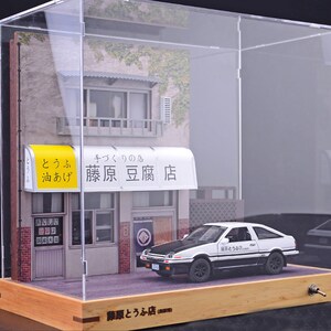 May include: A white and black toy car with the Japanese text "藤原豆腐店" on the side, sitting in front of a miniature storefront with a yellow awning that reads "藤原豆腐店" in Japanese. The storefront is inside a clear acrylic display case with a wooden base.