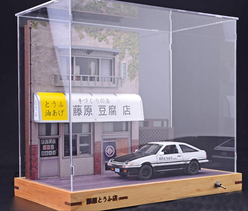 May include: A white and black toy car with the Japanese text "藤原豆腐店" on the side, sitting in front of a miniature storefront with a yellow awning that reads "藤原豆腐店" in Japanese. The storefront is inside a clear acrylic display case with a wooden base.