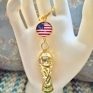 May include: A gold-toned keychain featuring a circular charm with the United States flag and a three-dimensional gold-colored trophy pendant. The trophy has a smooth, reflective finish with a green band detail at the base, attached by a metal ring.