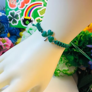 May include: A green string bracelet with a shamrock charm. The charm is made of metal and has a green enamel finish.