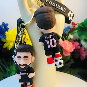 May include: Black and gold keychain with a 3D figurine of a soccer player wearing a black jersey with the number 10 and the text "MESSI" on the front. The keychain also has a black strap with the word "Football" printed on it.