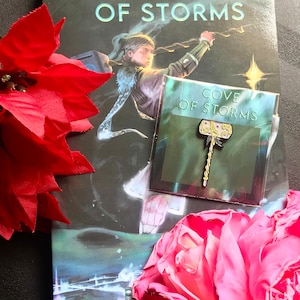 May include: A book titled "Cove of Storms" with a character on the cover holding a staff. A pin with the same title and a hammer design is on top of the book.