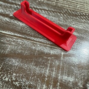 May include: A bright red, rectangular phone stand with a grooved channel and two raised end supports. The stand is made of a 3D-printed material and is designed to hold a phone or tablet. The stand is approximately 7 inches long.