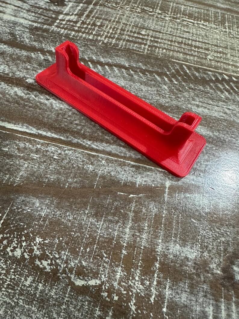May include: A bright red, rectangular phone stand with a grooved channel and two raised end supports. The stand is made of a 3D-printed material and is designed to hold a phone or tablet. The stand is approximately 7 inches long.